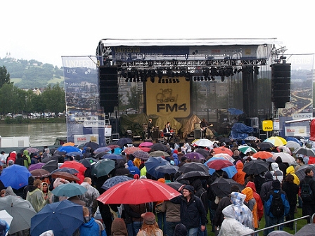 Das Publikum vor der FM4 Bühne. Es regnet. Im Hintergrund ist die Bühne mit der Band Kreisky zu erkennen. DAhinter die Donau. 